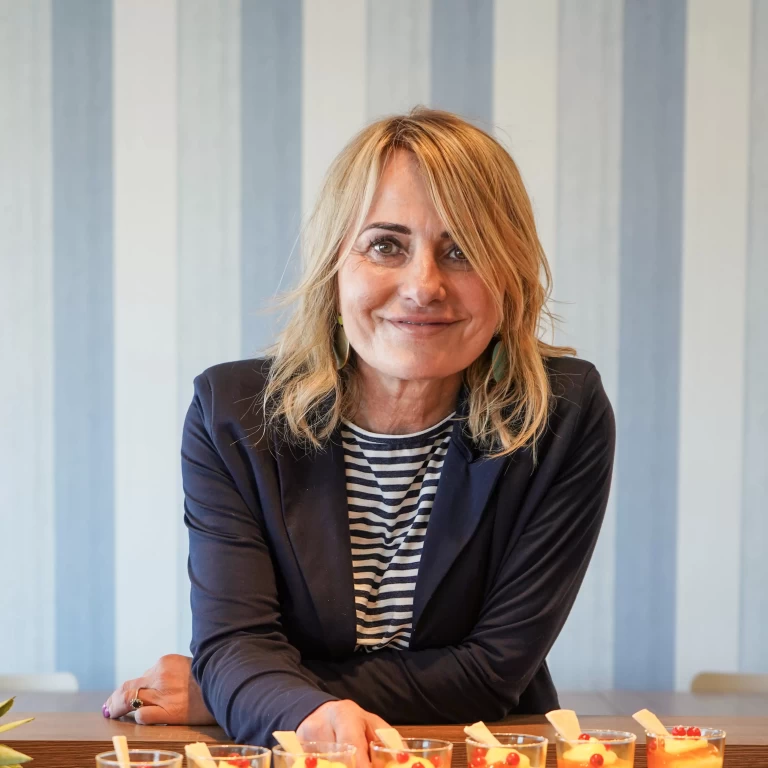 Donna sorridente con capelli biondi, indossa una giacca scura e maglia a righe, davanti a diversi bicchieri di dessert su un tavolo.