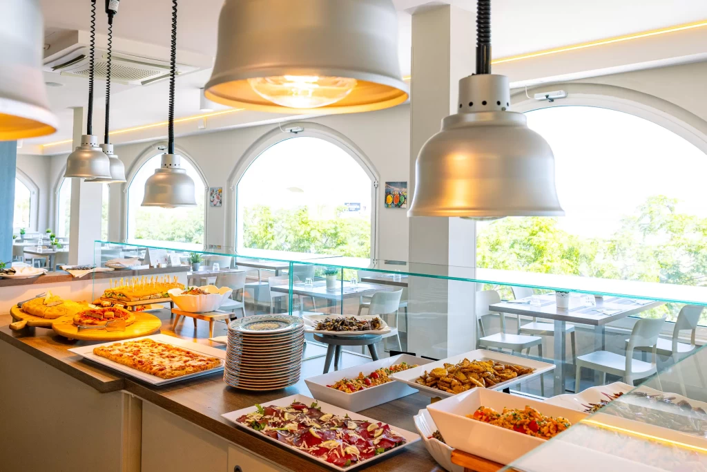 Buffet di un ristorante luminoso con tavoli apparecchiati in sottofondo.