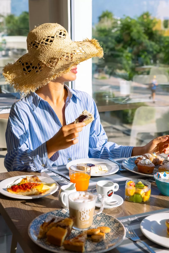 Donna con cappello a colazione, circondata da piatti dolci e salati in un ambiente luminoso.
