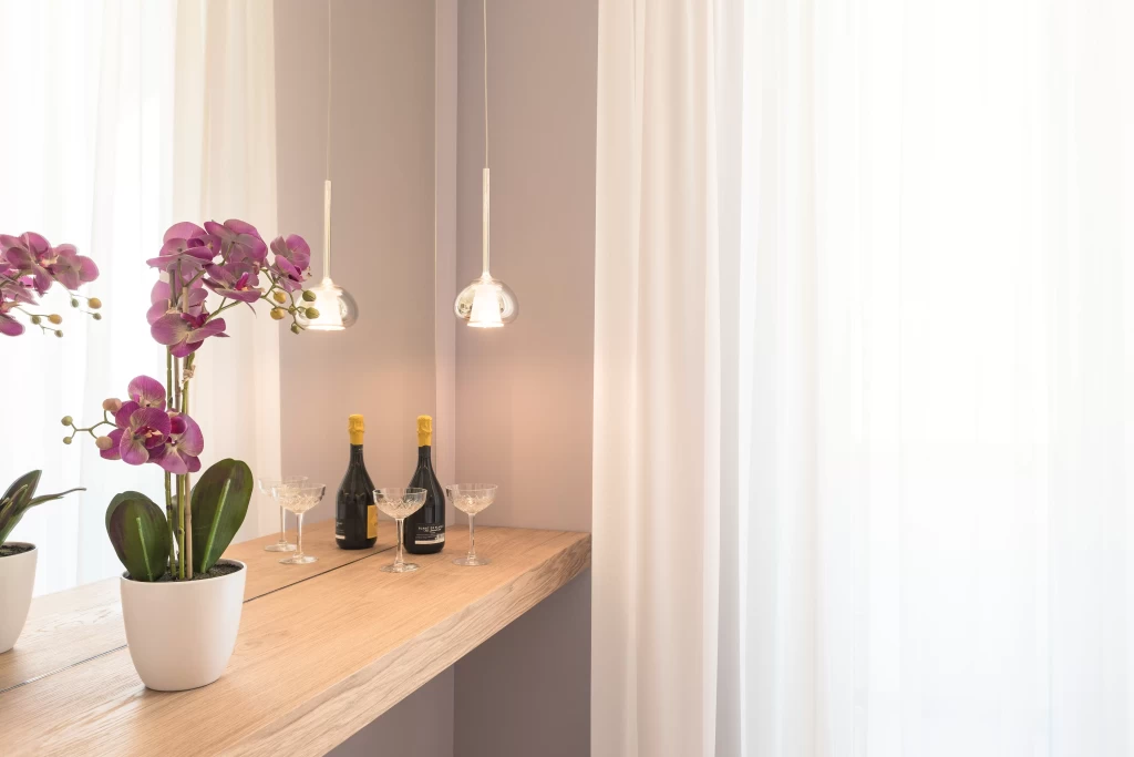 Piano d'appoggio con orchidee viola, bottiglie di vino e calici, illuminato da lampade pendenti.