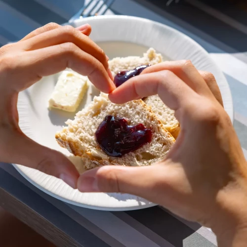 Mani formano un cuore attorno a una fetta di pane con marmellata su un piatto.