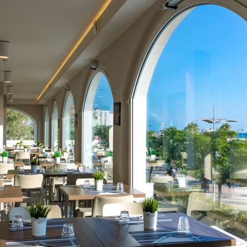 Interno di un ristorante luminoso con vista sul mare attraverso grandi finestre ad arco.