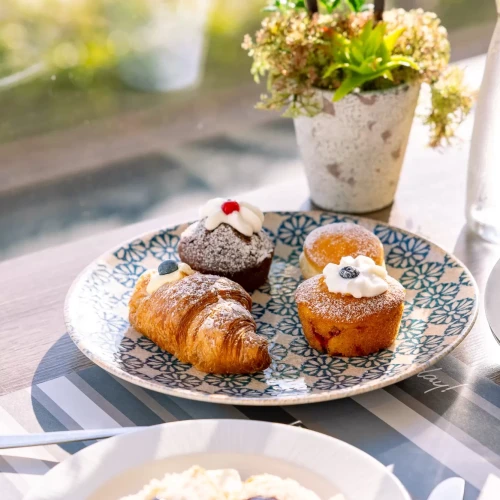 Piatto con croissant e muffin decorati, accanto a pane con marmellata e piantina su un tavolo luminoso.