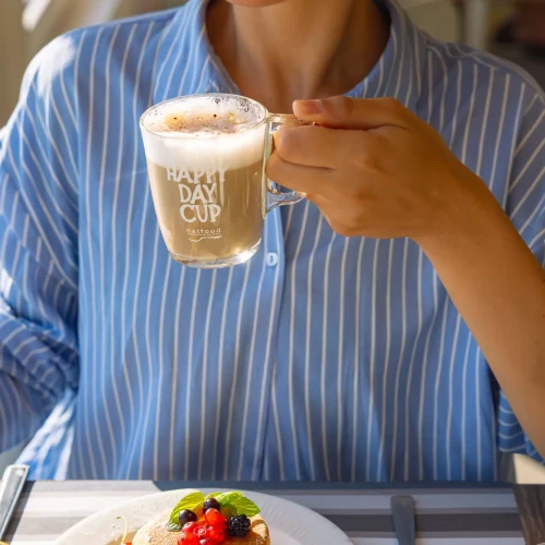 Donna con cappello di paglia sorseggia un cappuccino accanto a pancake con frutti di bosco.